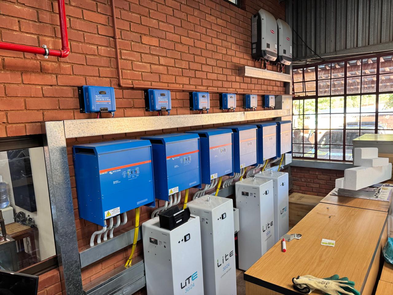 A commercial three-phase solar backup system featuring a wall-mounted bank of blue Victron Energy inverters, Victron MPPT charge controllers, Fronius grid-tied inverters, and four floor-standing white Freedom Won Lite 30/24 lithium batteries on a red face-brick wall.